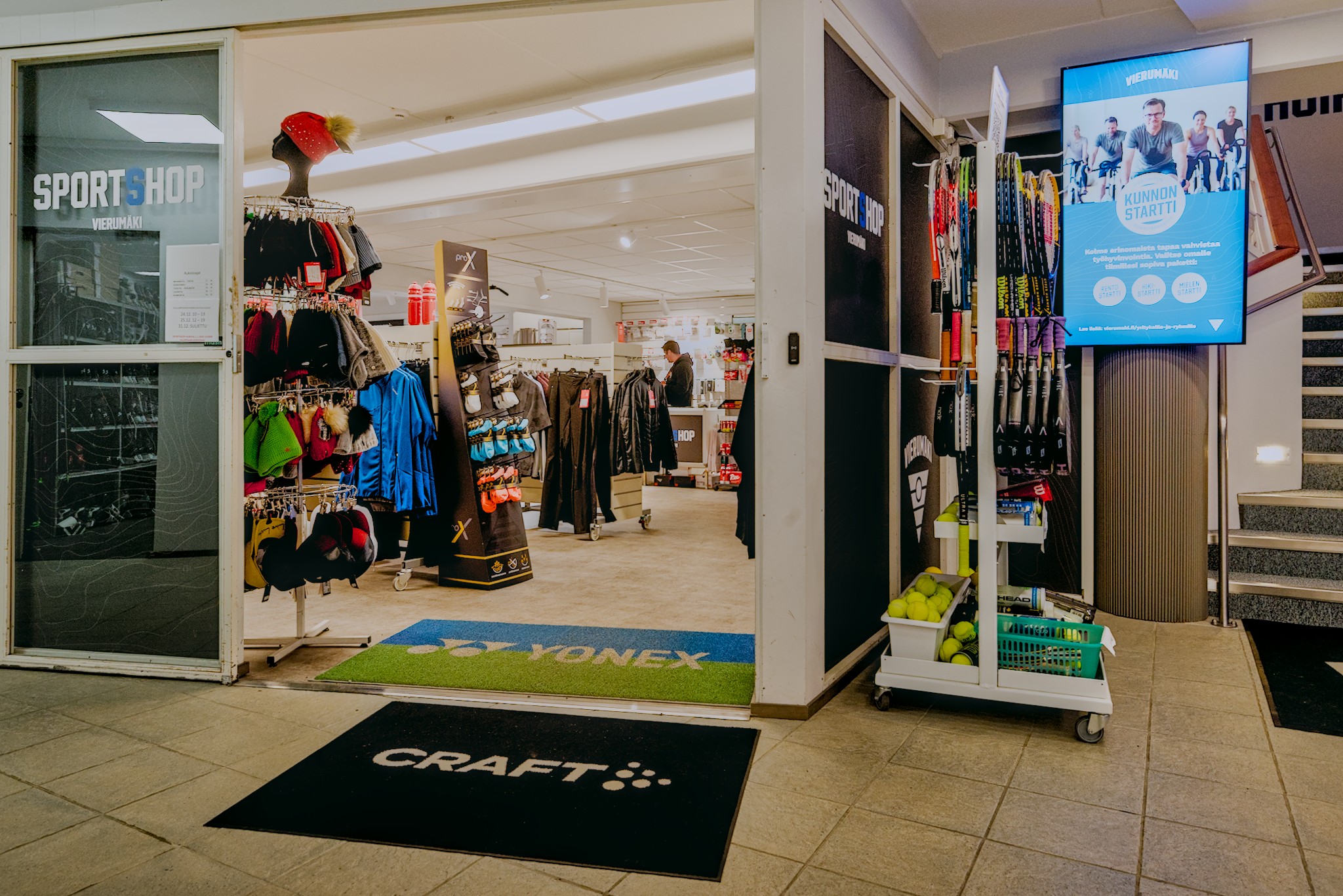 Sportshop Vierumäen valoisa myymälä tennis- ja padelhallin yhteydessä. Etualalla mailoja ja vaatteita, taustalla kauppias Toni Huttunen tiskin takana.